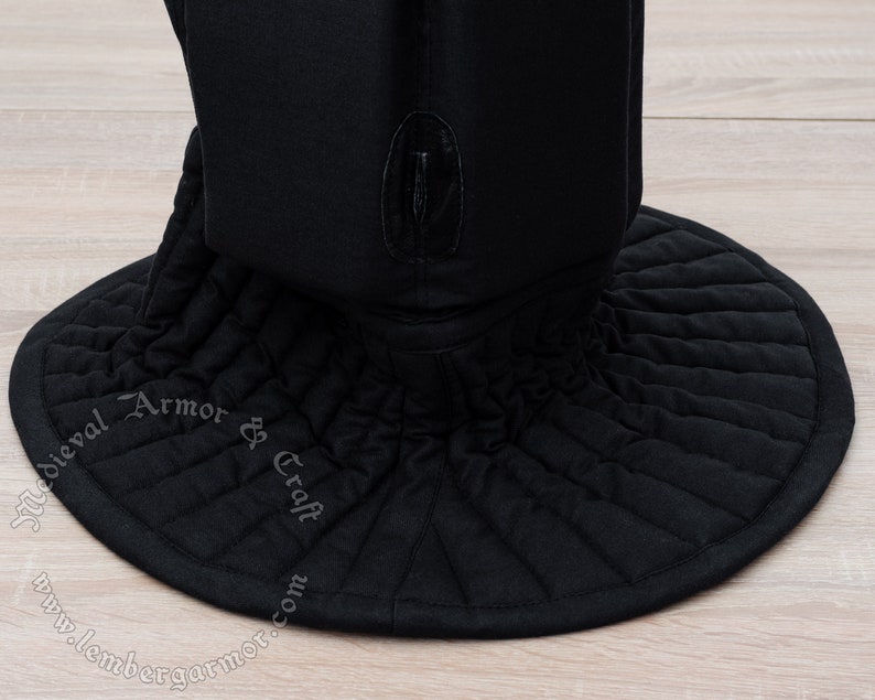 May include: Black quilted fabric medieval coif with a wide, round brim. The coif has a small, black leather tab on the top. The text "Medieval Armor & Craft" is embroidered on the brim.