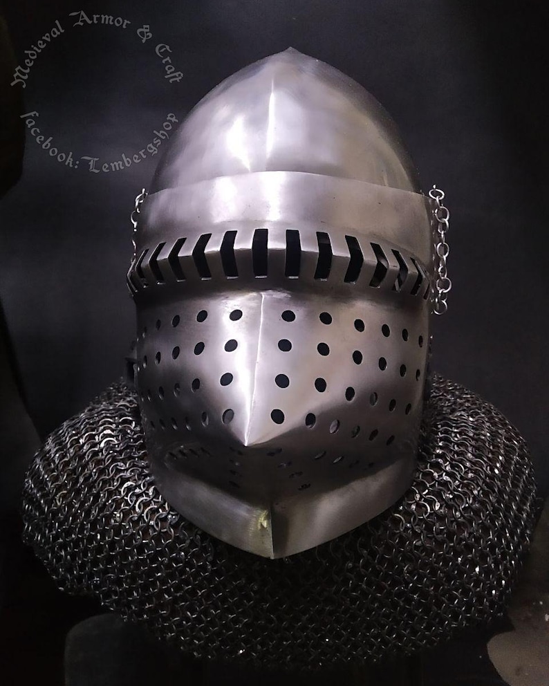 May include: A silver helmet with a chainmail coif. The helmet has a pointed top and a face mask with a series of small holes. The helmet is on display against a black background.