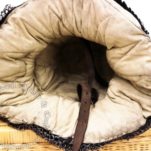 May include: The inside of a medieval helmet with a brown leather strap and a padded lining. The helmet is made of chainmail and has a  "Medieval Armor & Craft" logo.