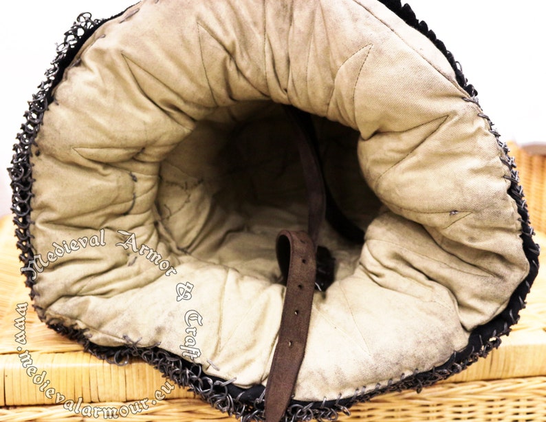 May include: The inside of a medieval helmet with a brown leather strap and a padded lining. The helmet is made of chainmail and has a  "Medieval Armor & Craft" logo.