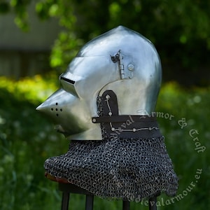 May include: A silver helmet with a pointed visor and a chainmail coif. The helmet has a hinged visor and a leather strap. The helmet is on a stand against a green background.