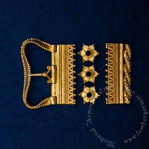 May include: A gold-colored belt buckle with a decorative design. The buckle has a hinged clasp and a decorative border. The buckle is made up of four separate pieces.