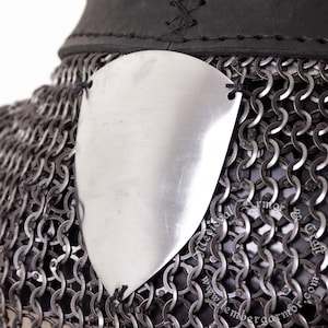 May include: Close-up of a section of medieval armor. The image shows a shiny, silver metal plate attached to a chainmail shirt. The chainmail is made of small, interlocking metal rings. A black leather collar is visible at the top.