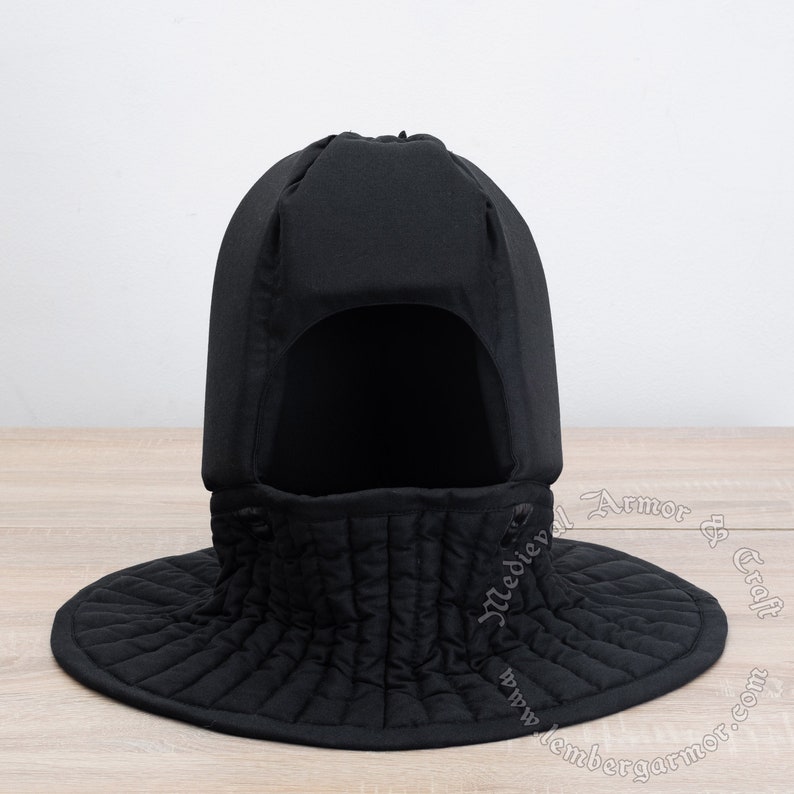 May include: Black quilted padded helmet with a neck guard and a face opening. The helmet is made of fabric and has a rounded shape.