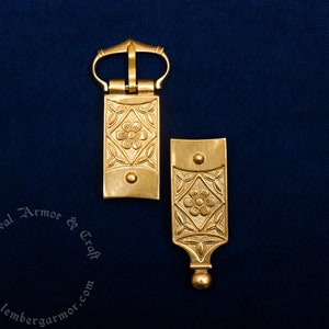 Puede incluir: Dos hebillas de cinturón de metal dorado con diseños florales y de hojas. Las hebillas son rectangulares con una flor en el centro y están sobre un fondo azul oscuro. Las hebillas son probablemente para recreaciones medievales o históricas.