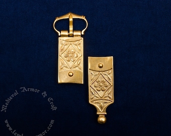 May include: Two gold-colored metal belt buckles with floral and leaf designs. The buckles are rectangular with a flower in the center and are set against a dark blue background. The buckles are likely for medieval or historical reenactment.