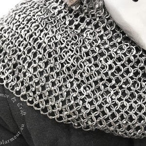 May include: A silver chainmail collar, a type of medieval armor, made of interlocking metal rings. The collar is shown on a dark fabric background.