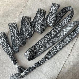 May include: A collection of handwoven belts and braids in grey and white, featuring geometric patterns. The textiles are displayed on a neutral linen background. The braids are finished with tassels. These accessories are suitable for craft projects.