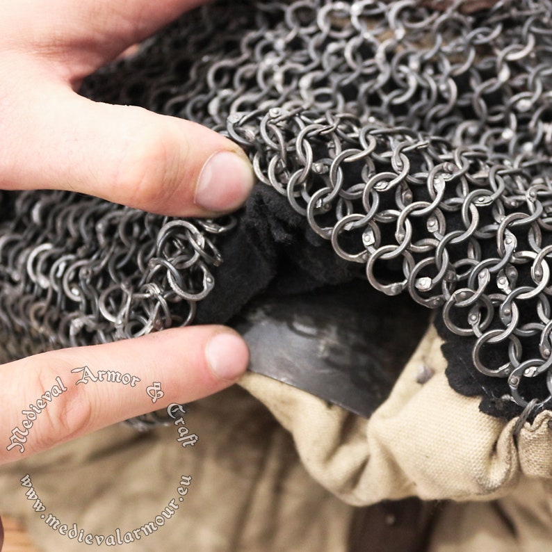 May include: Close-up of a chainmail coif with a black lining. The coif is made of interlocking metal rings and is folded over a piece of fabric. The fabric has the text "Medieval Armor & Craft www.medievalarmour.com" printed on it.