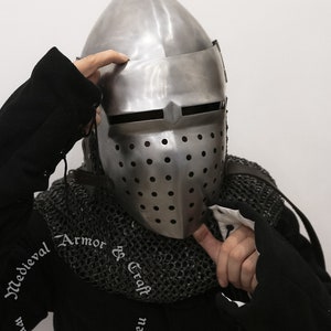 May include: A silver helmet with a pointed top and a face mask with holes. The helmet is worn by a person wearing a black shirt and chainmail.