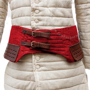 May include: Red quilted belt with brown leather straps and buckles. The belt has two rows of stitching and two rows of holes on each side. The belt is worn over a white linen garment.