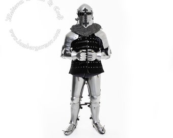 May include: A full suit of medieval armour in silver with a black under tunic. The armour includes a helmet with a cross on the front, gauntlets, and leg armour. The armour is standing on a white background.