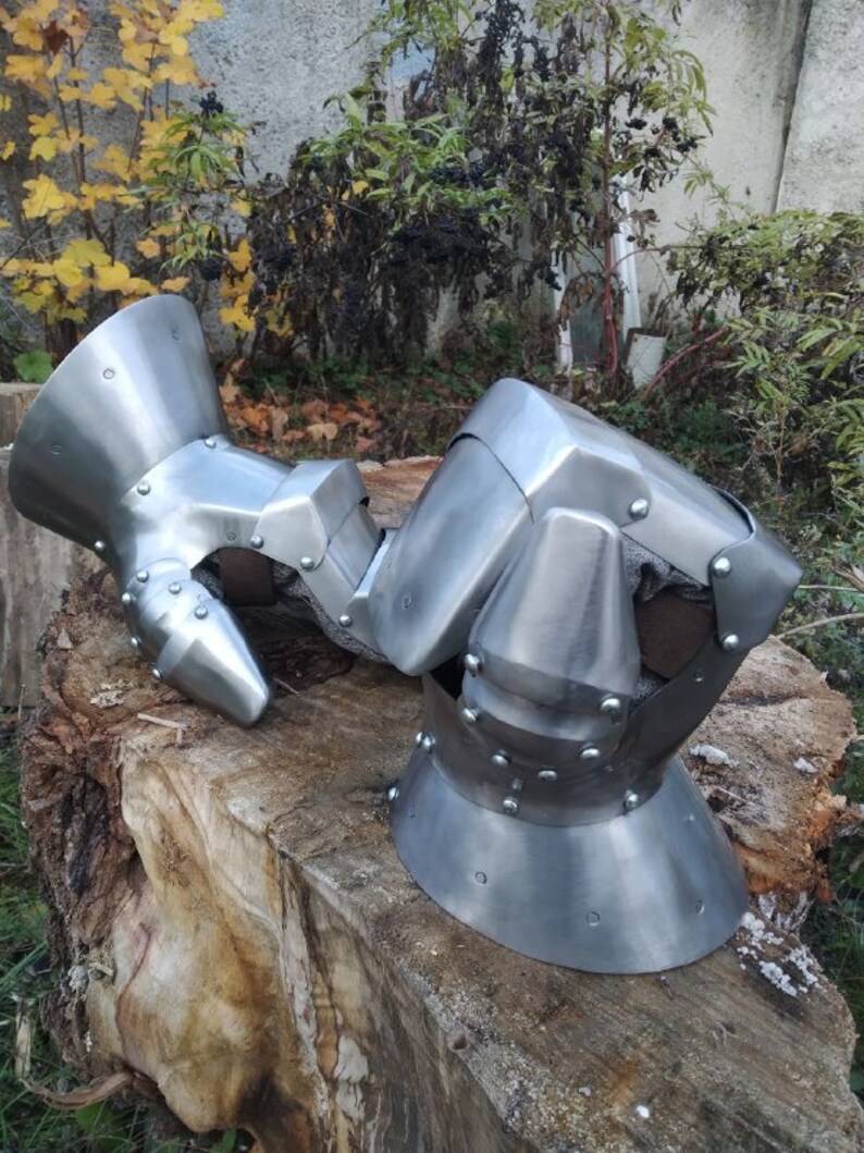 K&ouml;nnte beinhalten: Ein Paar mittelalterliche Handschuhe aus Metall mit silberner Oberfl&auml;che. Die Handschuhe sind so konzipiert, dass sie die H&auml;nde und Finger im Kampf sch&uuml;tzen. Die Handschuhe bestehen aus mehreren Metallplatten, die miteinander vernietet sind. Die Handschuhe haben eine spitze Form an den Kn&ouml;cheln und eine abgerundete Form auf der R&uuml;ckseite der Hand.