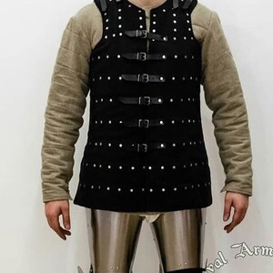 May include: A black leather vest with a studded design and multiple straps and buckles. The vest is worn over a long-sleeved shirt.