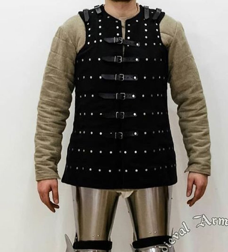 May include: A black leather vest with a studded design and multiple straps and buckles. The vest is worn over a long-sleeved shirt.