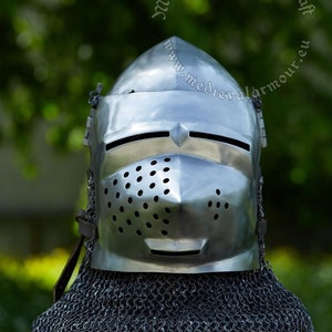 May include: A silver helmet with a chainmail coif, featuring a visor with a small opening for the eyes and a perforated nose guard. The helmet is on a wooden stand with a red cushion.