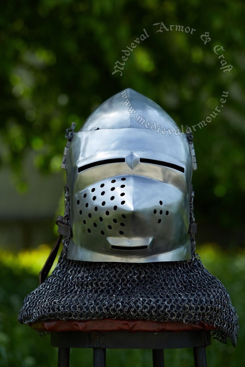 May include: A silver helmet with a chainmail coif, featuring a visor with a small opening for the eyes and a perforated nose guard. The helmet is on a wooden stand with a red cushion.