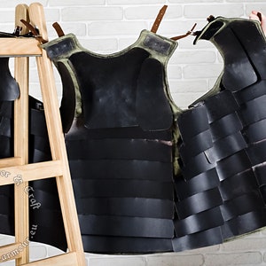 May include: A black and green leather armour vest with straps and buckles. The vest is made of overlapping plates and is hanging on a wooden rack.