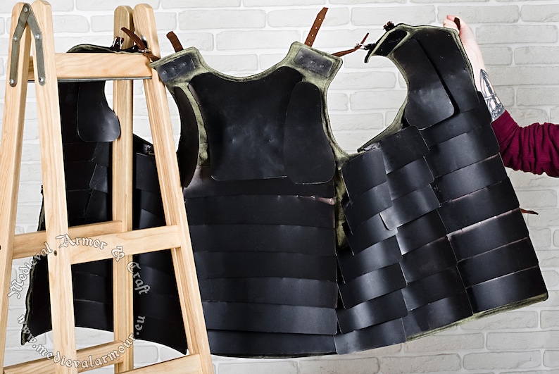 May include: A black and green leather armour vest with straps and buckles. The vest is made of overlapping plates and is hanging on a wooden rack.