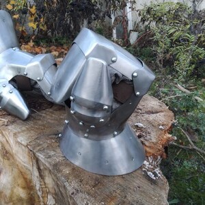 K&ouml;nnte beinhalten: Nahaufnahme eines silbernen Metallhelms mit spitzer Spitze und abgerundetem Gesichtschutz. Der Helm hat Nieten und eine strukturierte Oberfl&auml;che. Der Helm liegt auf einem St&uuml;ck Holz.