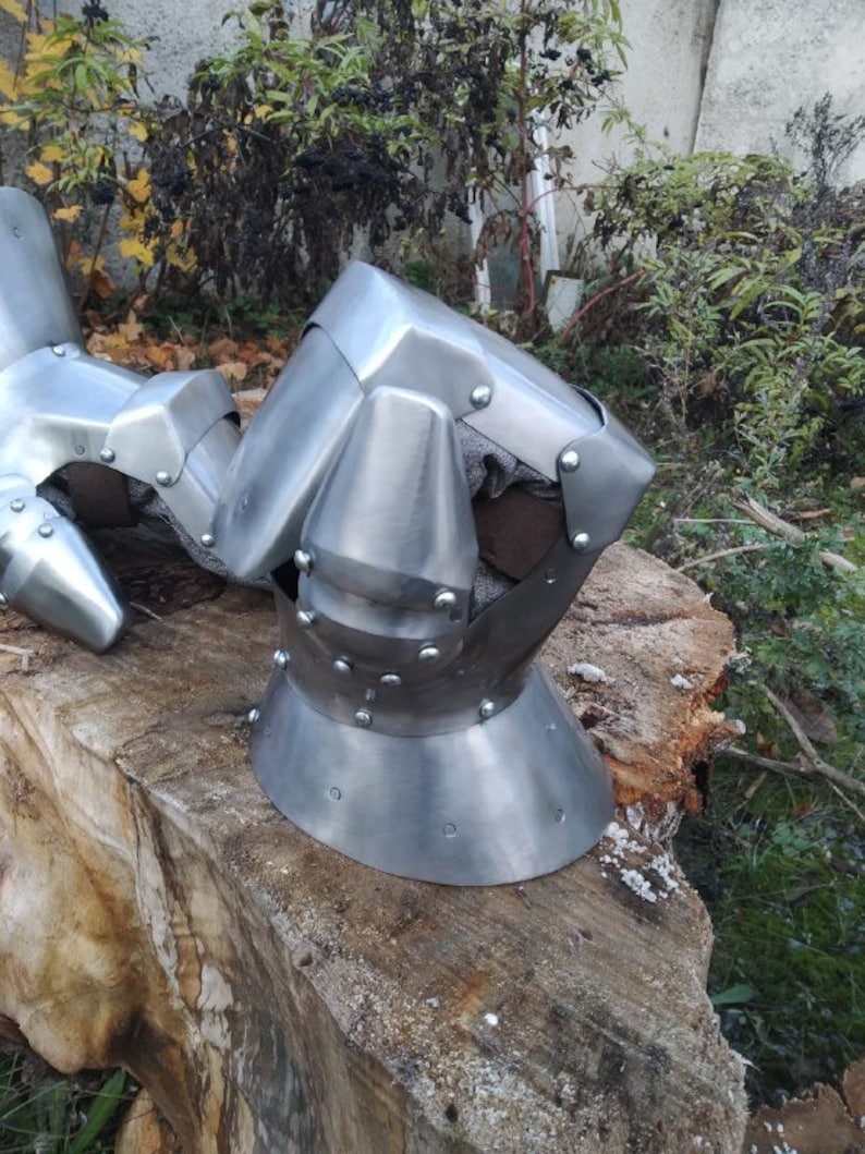 K&ouml;nnte beinhalten: Nahaufnahme eines silbernen Metallhelms mit spitzer Spitze und abgerundetem Gesichtschutz. Der Helm hat Nieten und eine strukturierte Oberfl&auml;che. Der Helm liegt auf einem St&uuml;ck Holz.