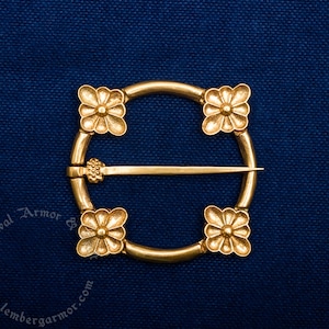 Pode incluir: Um broche circular dourado com quatro acentos decorativos em forma de flor. O broche tem um fecho de pino.