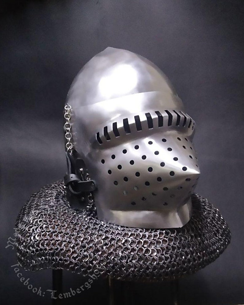 May include: A silver helmet with a face mask and a chainmail coif. The helmet has a pointed nose guard and a series of small holes in the face mask. The chainmail coif is made of small metal rings.