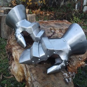 K&ouml;nnte beinhalten: Ein Paar silberne Metallhandschuhe mit beweglichen Fingern und einer Schutzmanschette am Handgelenk. Die Handschuhe liegen auf einem Baumstumpf.