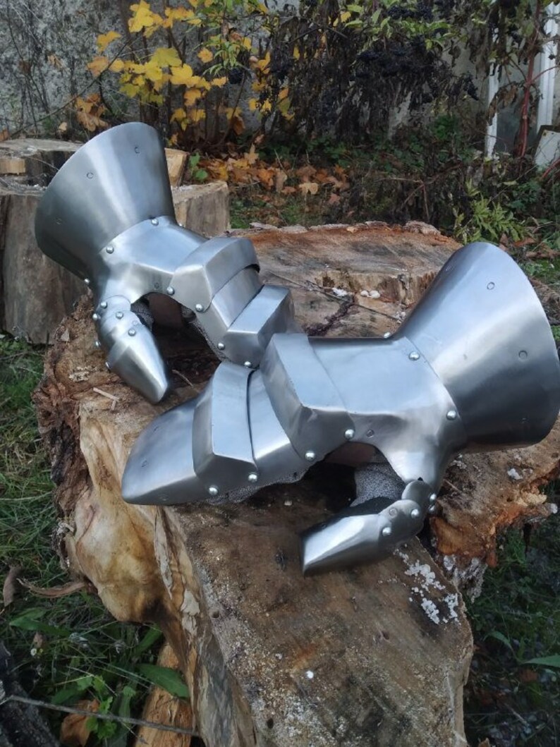 K&ouml;nnte beinhalten: Ein Paar silberne Metallhandschuhe mit beweglichen Fingern und einer Schutzmanschette am Handgelenk. Die Handschuhe liegen auf einem Baumstumpf.