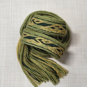 May include: A handwoven green and gold belt with a geometric pattern. The belt is rolled up, with a long fringe of loose threads at the end. The design features dark green and gold woven bands.