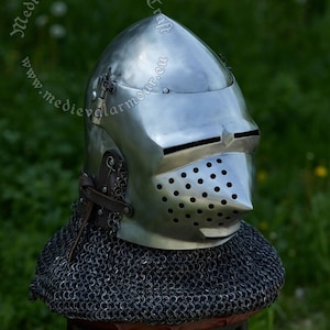 May include: A silver helmet with a pointed visor and a chainmail coif. The helmet has a series of small holes in the visor and is sitting on a wooden stool.
