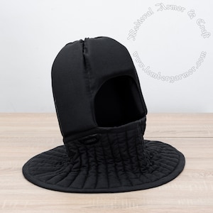 May include: Black padded medieval coif with a neck guard. The coif has a hole for the face and a quilted neck guard. The coif is on a wooden surface.