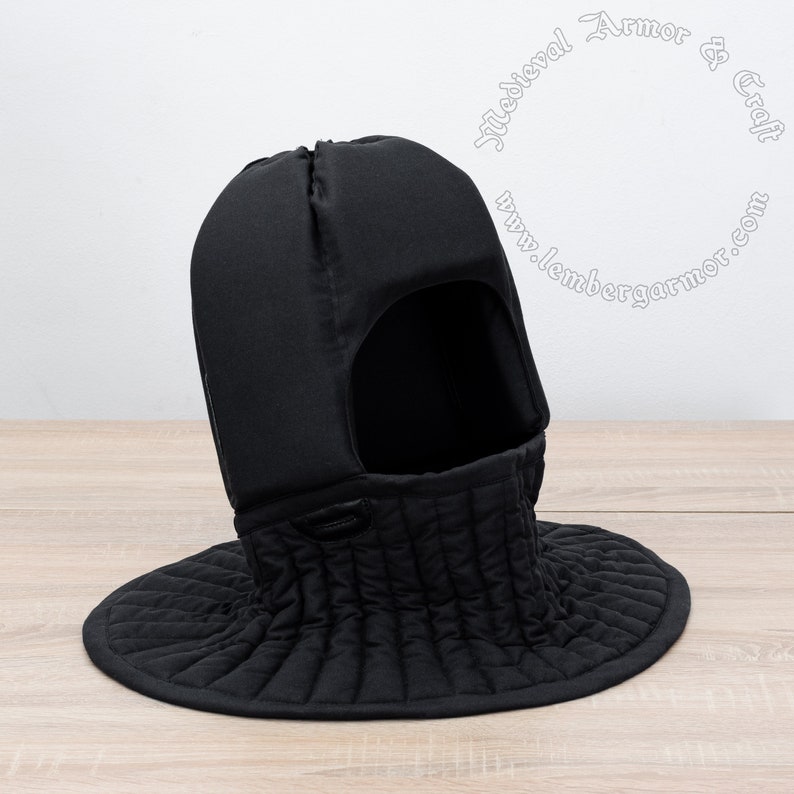 May include: Black padded medieval coif with a neck guard. The coif has a hole for the face and a quilted neck guard. The coif is on a wooden surface.