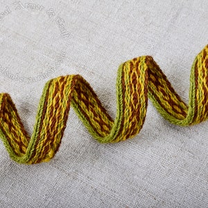 May include: A close-up of a woven green and brown ribbon. The ribbon is twisted and curled on a light gray fabric background. The text "Medieval Armor & Craft" is visible in the background.