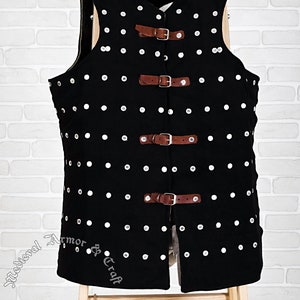 May include: Black padded vest with silver rivets and brown leather straps and buckles. The vest is displayed on a wooden stool against a white brick wall.