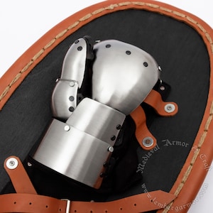 May include: A close-up of a medieval gauntlet made of polished steel, with articulated fingers and a cuff. The gauntlet is set against a black background, with a brown leather border and straps.