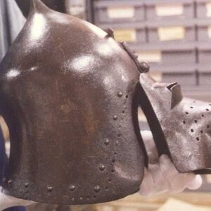 May include: A close-up of a brown metal helmet with a pointed nose guard and a textured surface. The helmet has a series of small rivets around the edges.