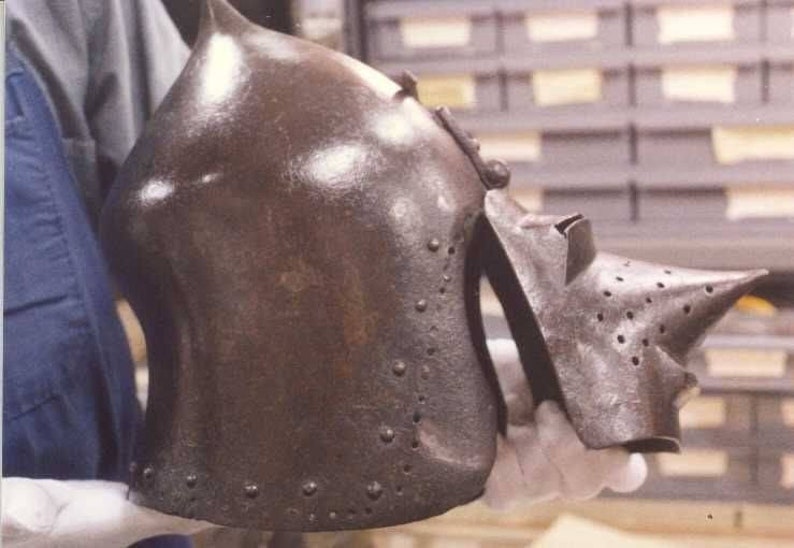 May include: A close-up of a brown metal helmet with a pointed nose guard and a textured surface. The helmet has a series of small rivets around the edges.