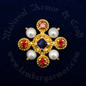May include: A gold cross pendant with a blue gemstone in the centre and four red gemstones around it. The cross is surrounded by four white pearls.