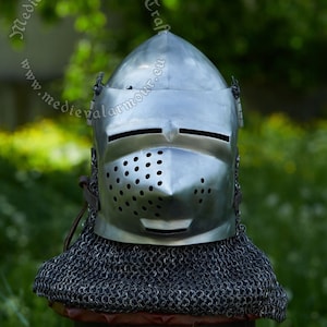 May include: A silver helmet with a visor and a chainmail coif. The helmet has a series of small holes in the visor. The helmet is sitting on a wooden stool.