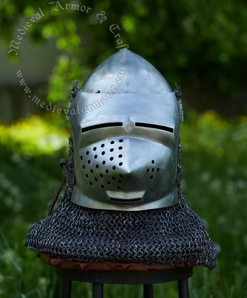 May include: A silver helmet with a visor and a chainmail coif. The helmet has a series of small holes in the visor. The helmet is sitting on a wooden stool.