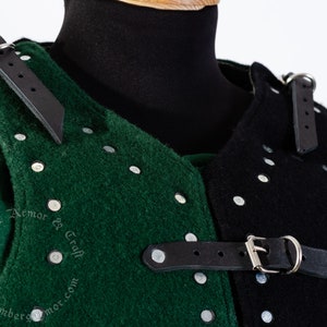 May include: Close-up of a green and black padded armor with metal rivets and leather straps. The armor is made of thick fabric and has a medieval design.