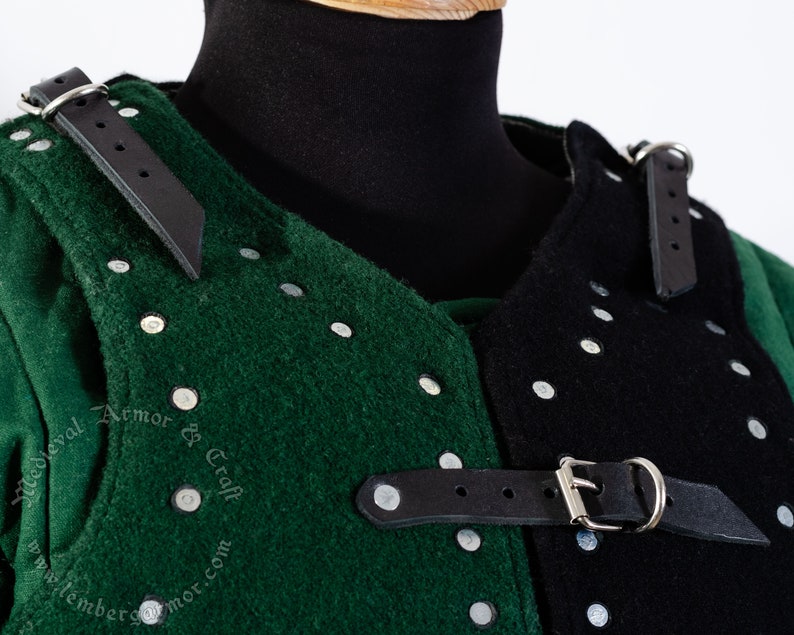 May include: Close-up of a green and black padded armor with metal rivets and leather straps. The armor is made of thick fabric and has a medieval design.