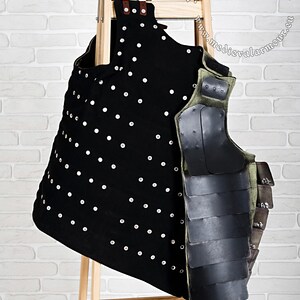 May include: A black and green padded armour vest with metal plates and rivets. The vest is hanging on a wooden rack against a white brick wall. The text "Medieval Armor & Craft" is visible in the upper right corner of the image.