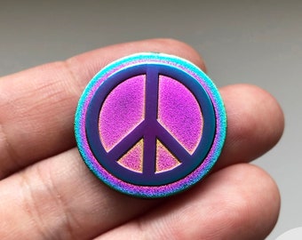 May include: A round, iridescent peace sign pin with a purple center and a gold outline. The pin is held in a hand.