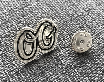 May include: Silver-toned metal pin with a black outline of the letters "OG". The pin has a silver-toned back with a circular design.