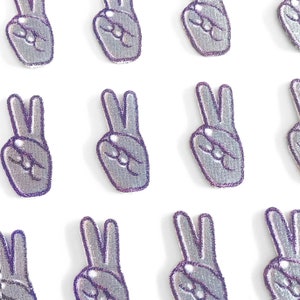 May include: A collection of silver peace sign patches with a purple glitter outline. The patches are embroidered with a detailed hand gesture, showing the index and middle fingers raised. The patches are arranged in a grid pattern against a white background.