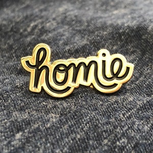 May include: A gold-colored enamel pin with the word "homie" in black script. The pin has a glossy finish and is set against a denim background. The word "homie" is written in a cursive font.