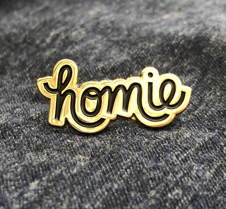 May include: A gold-colored enamel pin with the word "homie" in black script. The pin has a glossy finish and is set against a denim background. The word "homie" is written in a cursive font.