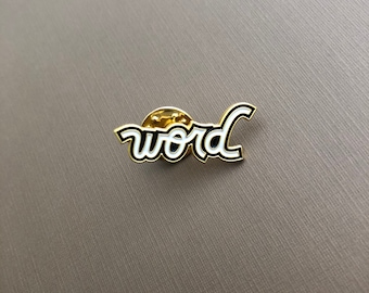 May include: A gold-colored enamel pin with the word "word" in a cursive font. The word is outlined in black with a white fill. The pin is on a textured, neutral-toned surface.